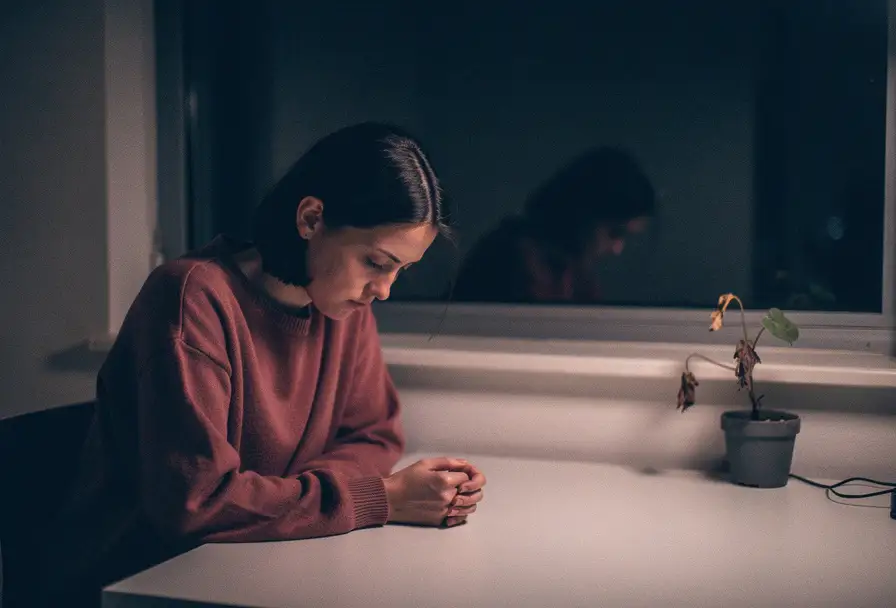 Uitgeputte vrouw alleen aan minimalistisch bureau, schouders gebogen, weerspiegeld in donker raam, verwelkte plant, gedempte roze en blauwe tinten.