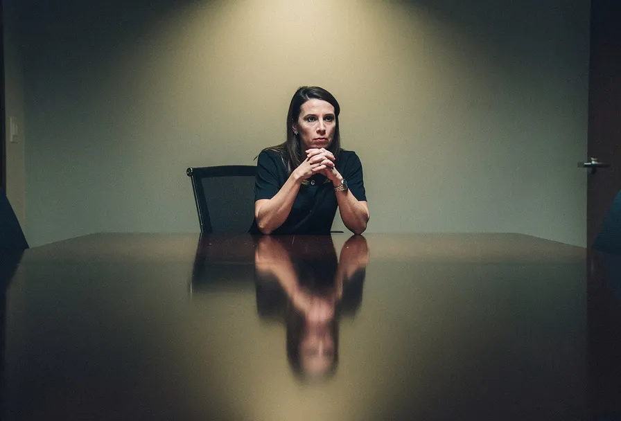 Vrouw met gevouwen handen aan vergadertafel, lege stoel tegenover haar, zacht amberkleurig licht in stille, gespannen sfeer.